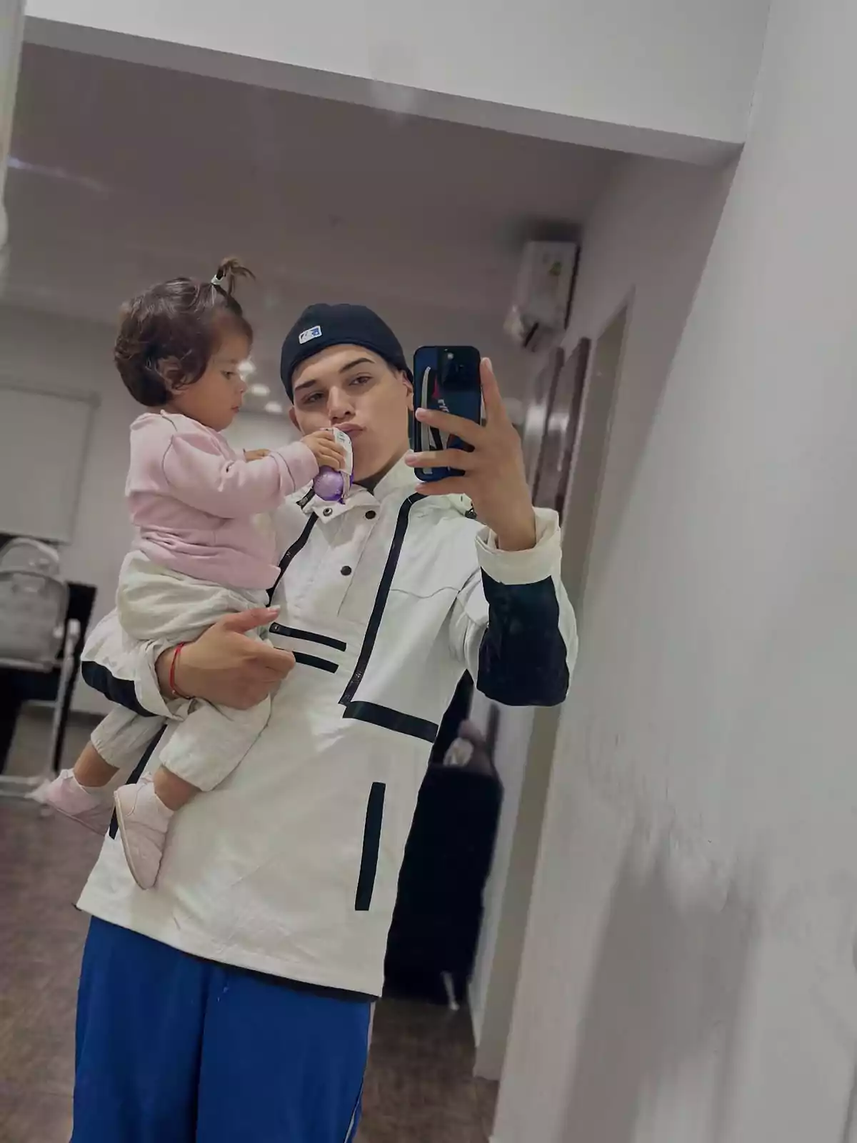 Un joven con gorra negra y chaqueta blanca sostiene a una niña pequeña vestida de rosa mientras ambos se miran en un espejo y él toma una selfie con su teléfono móvil Un joven con gorra negra y chaqueta blanca sostiene a una niña pequeña vestida de rosa mientras ambos se miran en un espejo y él toma una selfie con su teléfono móvil