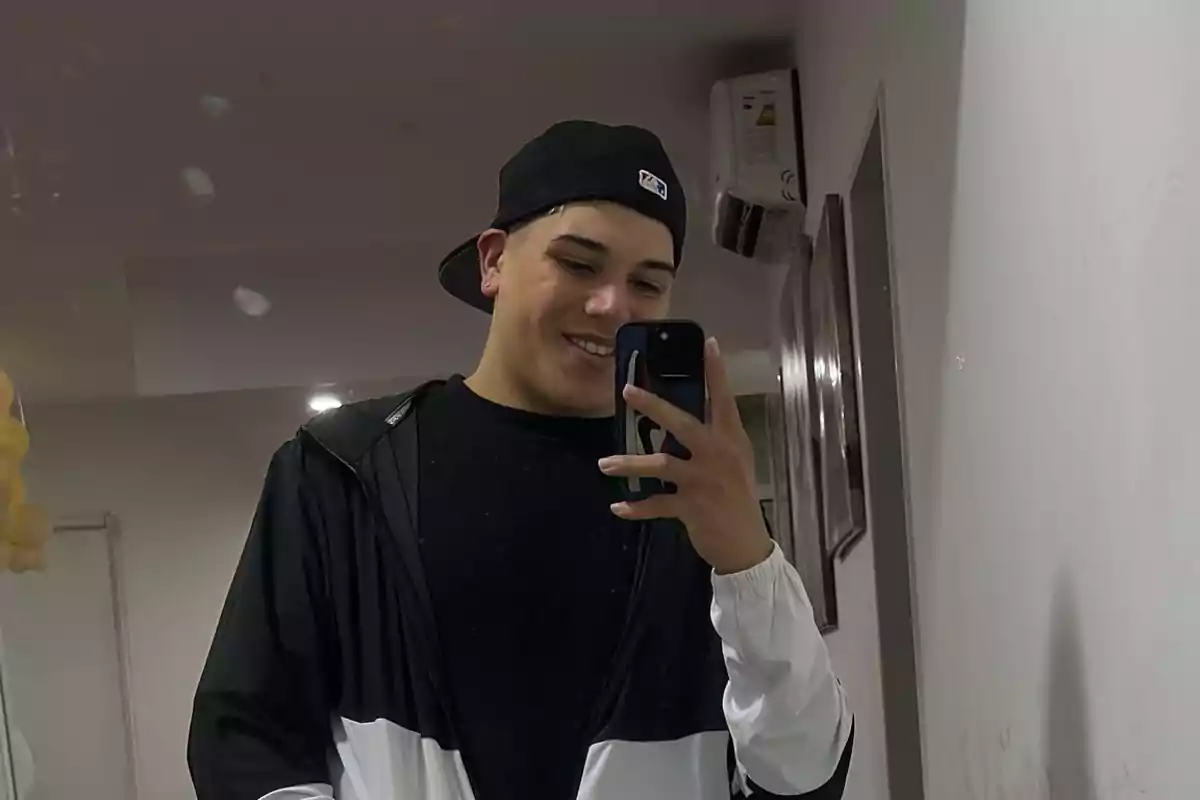 Joven con gorra negra hacia atrás tomándose una selfie frente a un espejo mientras sonríe, usando una chaqueta deportiva blanca y negra.