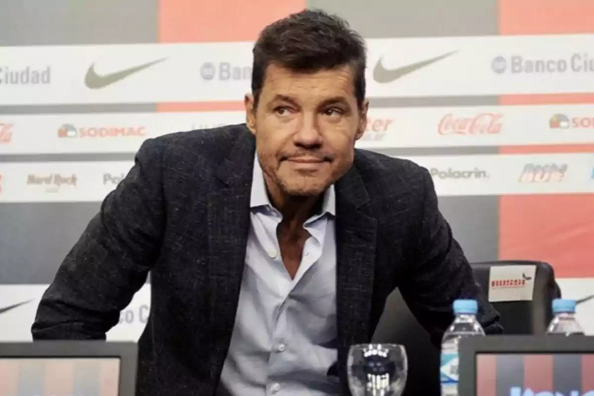 Hombre de cabello corto y barba incipiente con saco oscuro y camisa clara sentado frente a micrófonos en una conferencia de prensa con fondo de logotipos de marcas.
