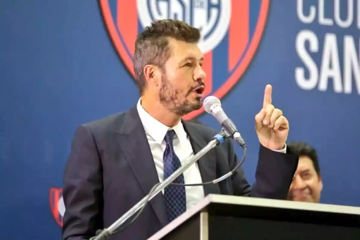 Hombre de traje hablando en un micrófono durante un evento en un club deportivo Hombre de traje hablando en un micrófono durante un evento en un club deportivo