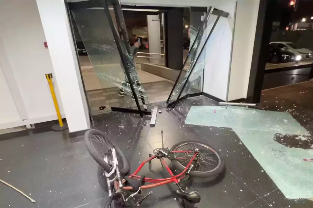 Bicicleta tirada en el suelo frente a una puerta de vidrio rota con fragmentos de cristal esparcidos en el piso dentro de un edificio Bicicleta tirada en el suelo frente a una puerta de vidrio rota con fragmentos de cristal esparcidos en el piso dentro de un edificio
