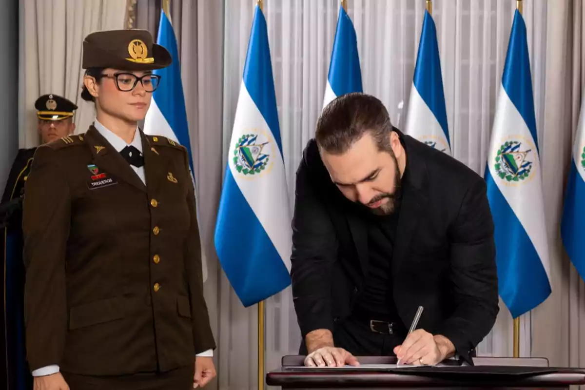 Un hombre vestido de negro firma un documento en una mesa mientras una mujer con uniforme militar lo observa, al fondo hay varias banderas de El Salvador