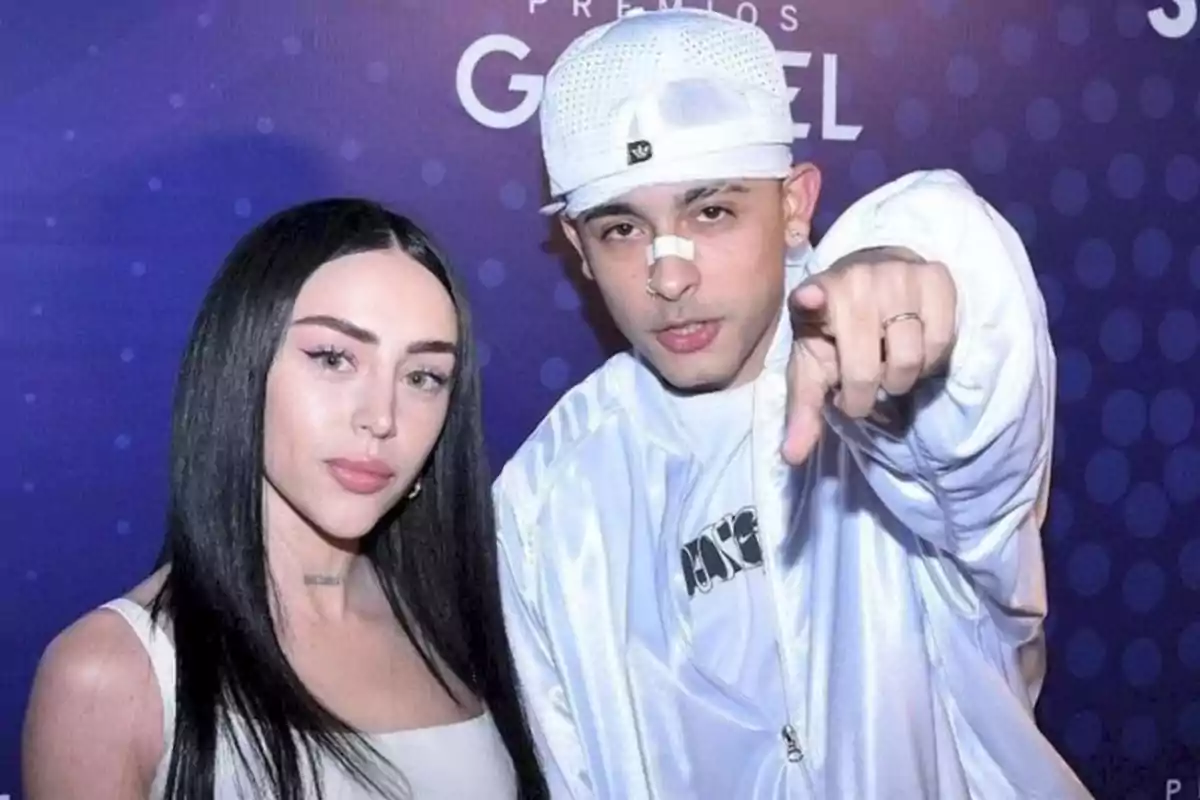 Two people pose in front of a blue background with dots and white letters: a woman with long, straight hair and a man wearing a white cap and a bandage on his nose points toward the camera. Two people pose in front of a blue background with dots and white letters: a woman with long, straight hair and a man wearing a white cap and a bandage on his nose points toward the camera.