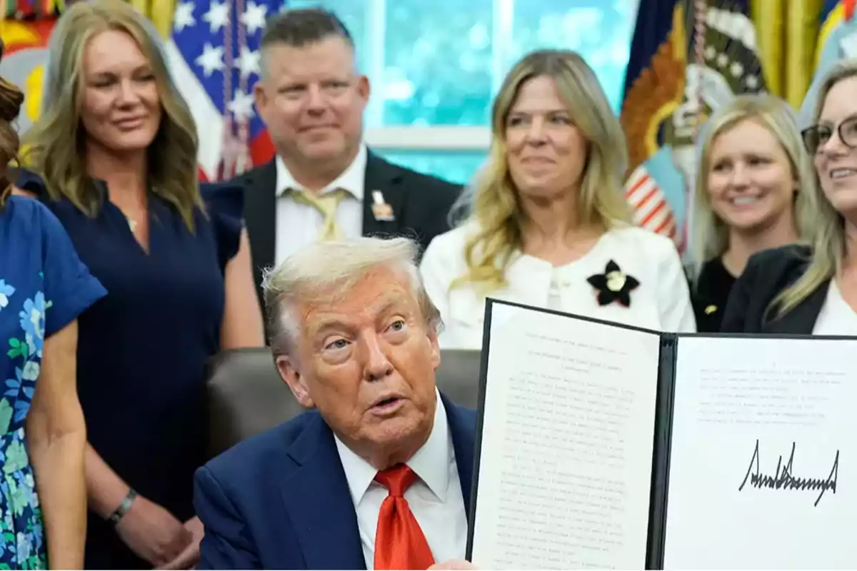 A blond-haired man in a blue suit holds a signed document while several people surround him and watch in an official office decorated with flags.