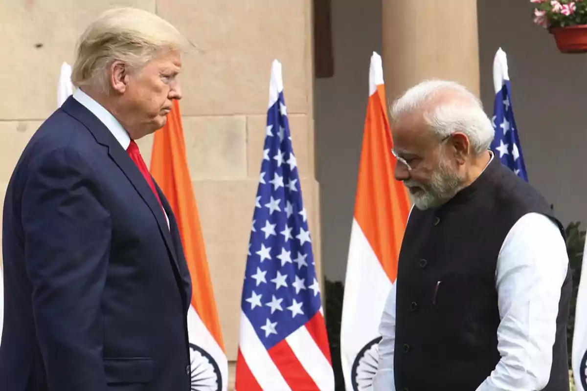 Two men in suits greet each other in front of United States and Indian flags in an official setting. Two men in suits greet each other in front of United States and Indian flags in an official setting.