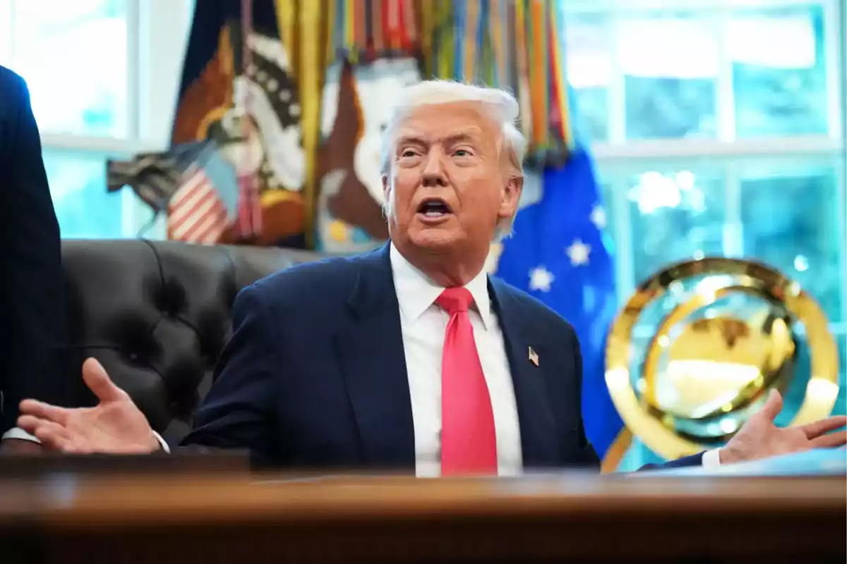 Blond-haired man in a dark suit and red tie sitting in an office with flags and decorations in the background