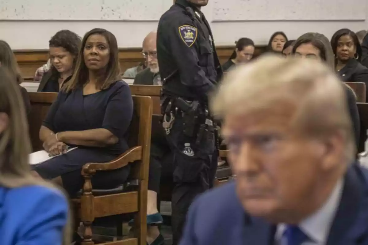 A woman sitting in a courtroom looks ahead while a police officer stands nearby and several people are seated in the background, with a blurry blond man in the foreground. A woman sitting in a courtroom looks ahead while a police officer stands nearby and several people are seated in the background, with a blurry blond man in the foreground.
