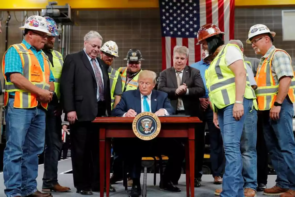 A man in a suit signs a document at an official desk surrounded by construction workers wearing vests and hard hats, while a United States flag hangs in the background. A man in a suit signs a document at an official desk surrounded by construction workers wearing vests and hard hats, while a United States flag hangs in the background.