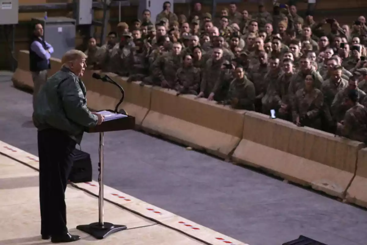 A man in a green jacket speaks at a podium in front of a large group of soldiers who watch him alertly in an enclosed space. A man in a green jacket speaks at a podium in front of a large group of soldiers who watch him alertly in an enclosed space.