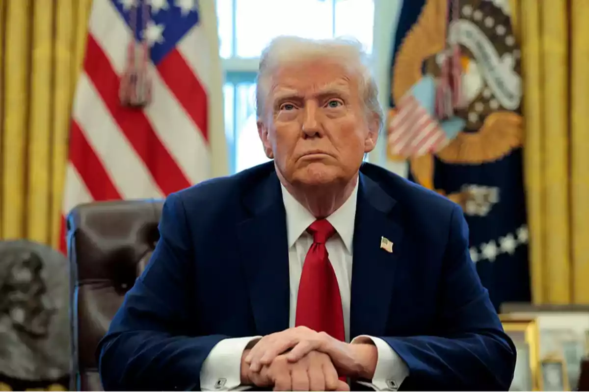 Hombre de cabello rubio con traje azul y corbata roja sentado en un escritorio con banderas de Estados Unidos al fondo