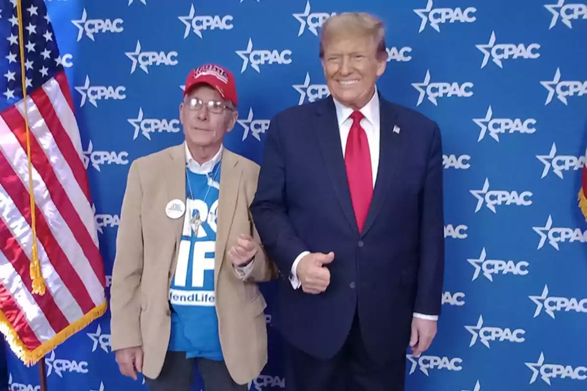 Dos hombres posan juntos frente a un fondo azul con logotipos de CPAC y una bandera de Estados Unidos a la izquierda uno de ellos lleva un traje oscuro con corbata roja y el otro viste una camiseta azul con mensaje, saco beige y gorra roja Dos hombres posan juntos frente a un fondo azul con logotipos de CPAC y una bandera de Estados Unidos a la izquierda uno de ellos lleva un traje oscuro con corbata roja y el otro viste una camiseta azul con mensaje, saco beige y gorra roja