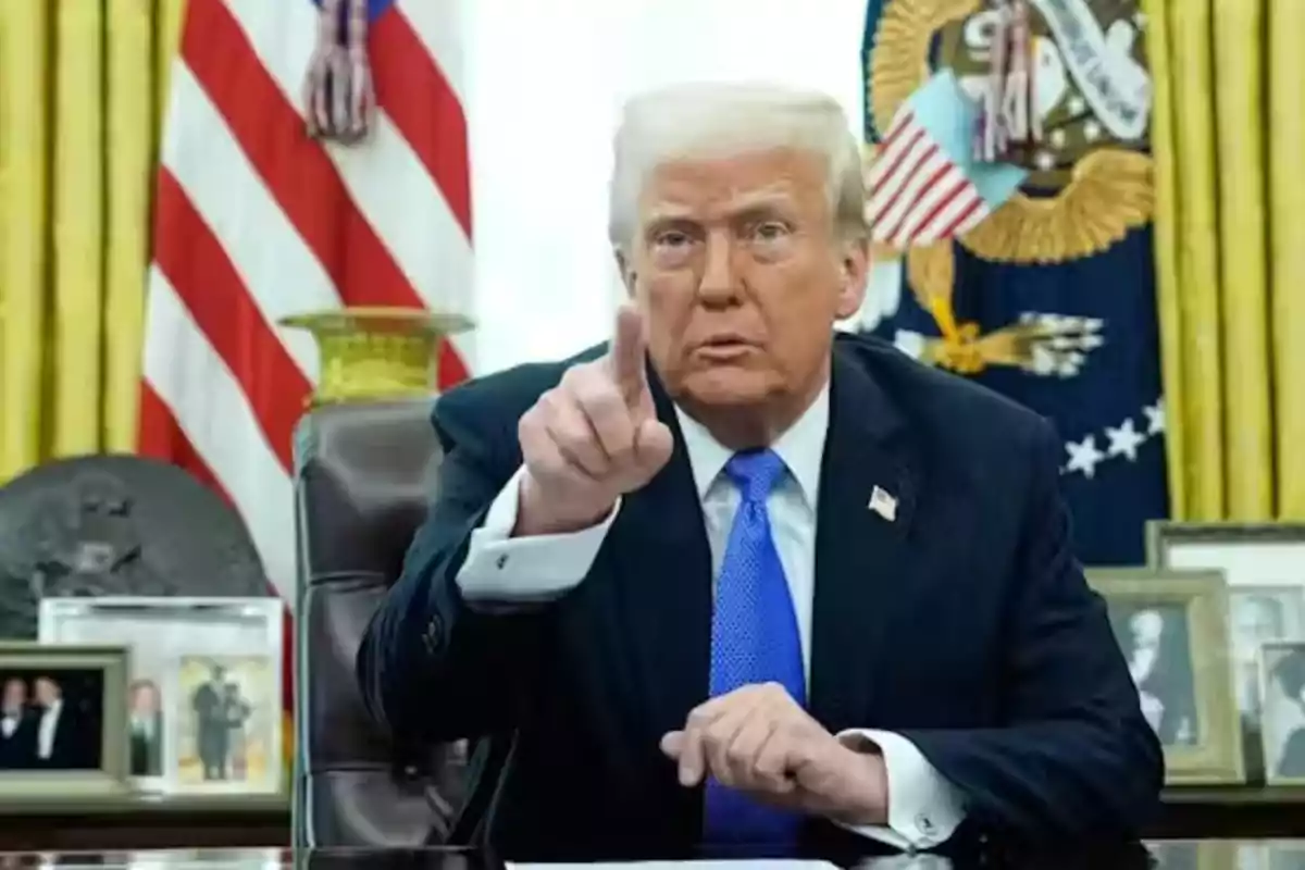 Hombre de cabello rubio con traje oscuro y corbata azul sentado en un escritorio señalando con el dedo índice hacia el frente con banderas y fotografías en el fondo