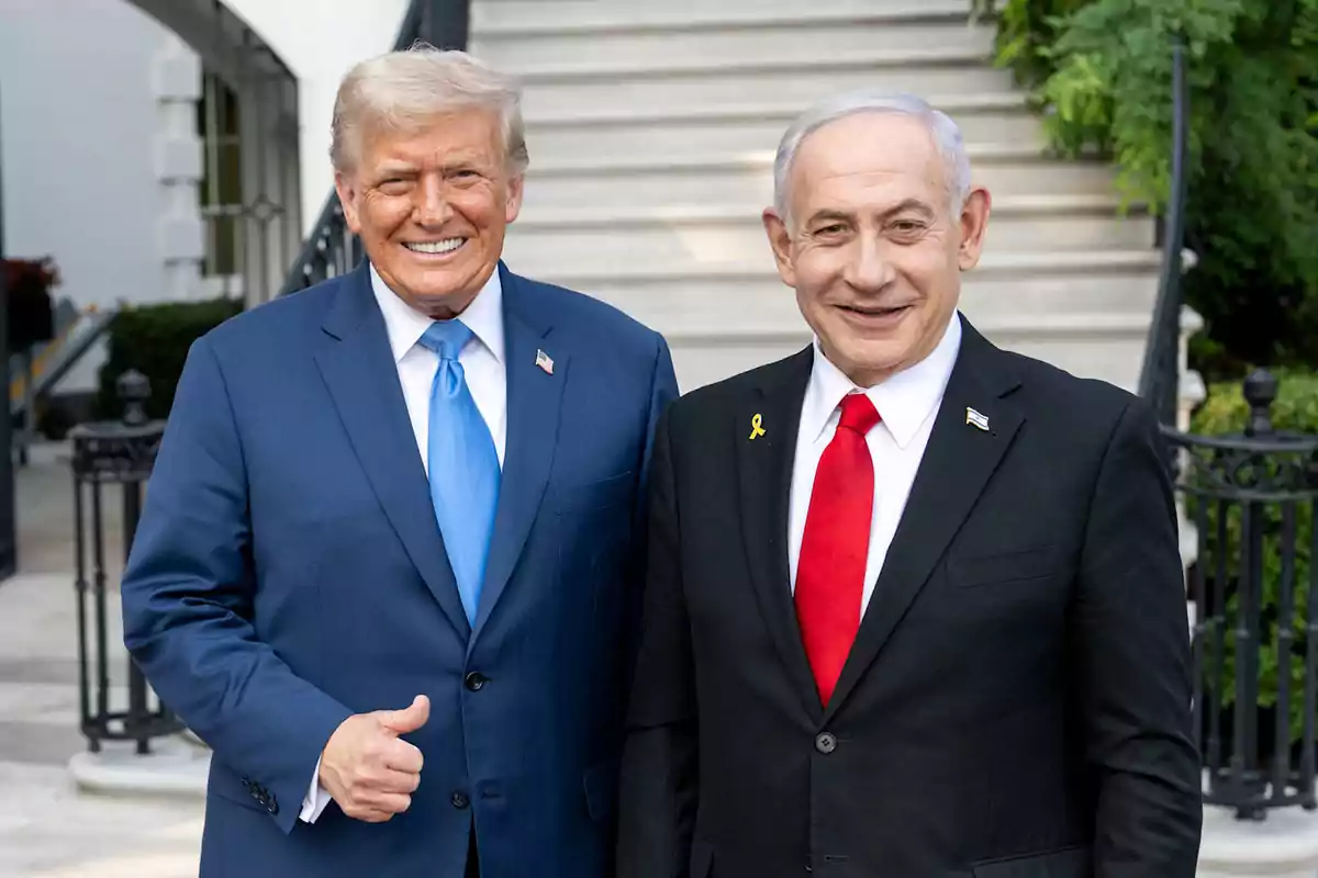 Two men in suits smile in front of an outdoor staircase, one wearing a blue tie and the other a red tie.