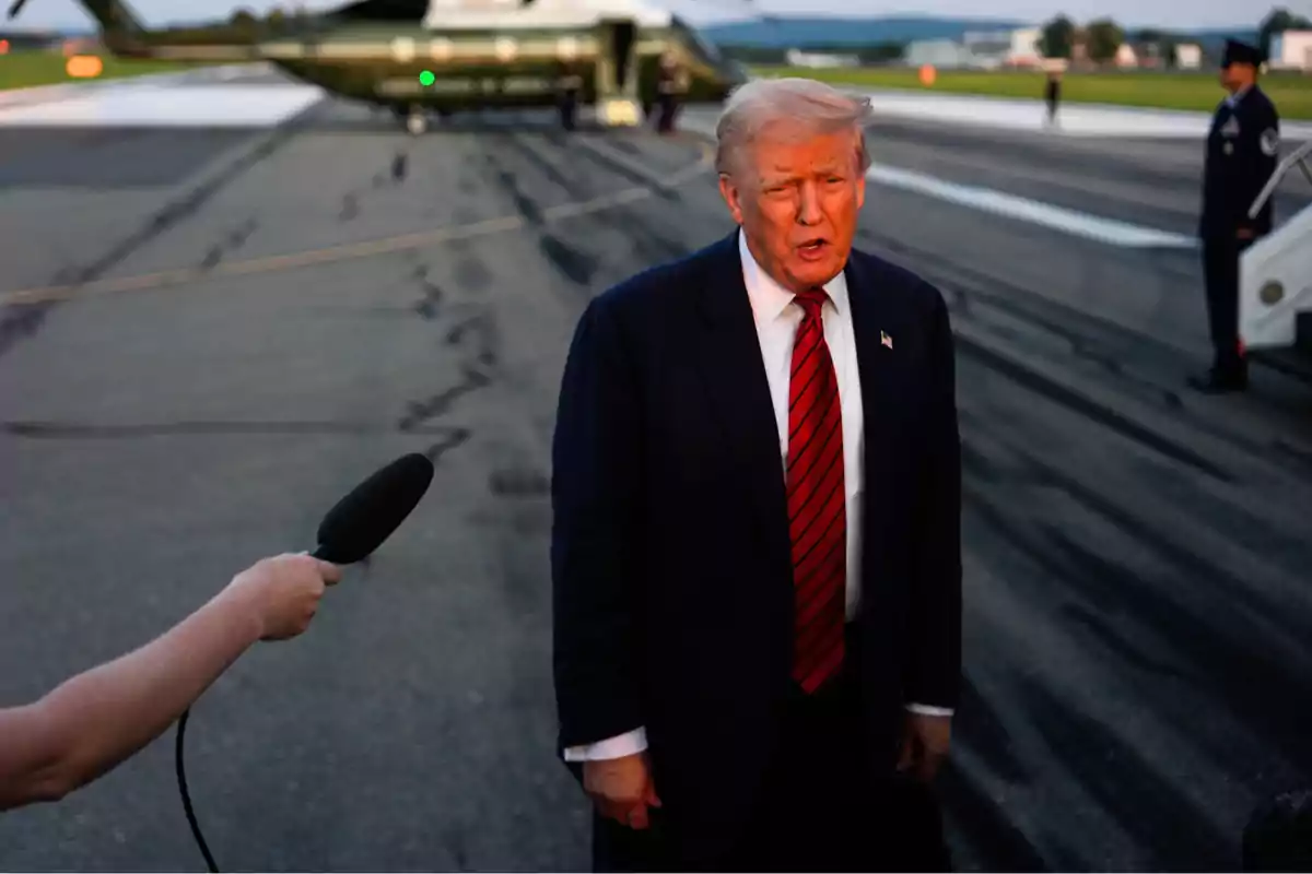 Hombre de cabello rubio con traje oscuro y corbata roja siendo entrevistado en una pista de aterrizaje con un helicóptero al fondo