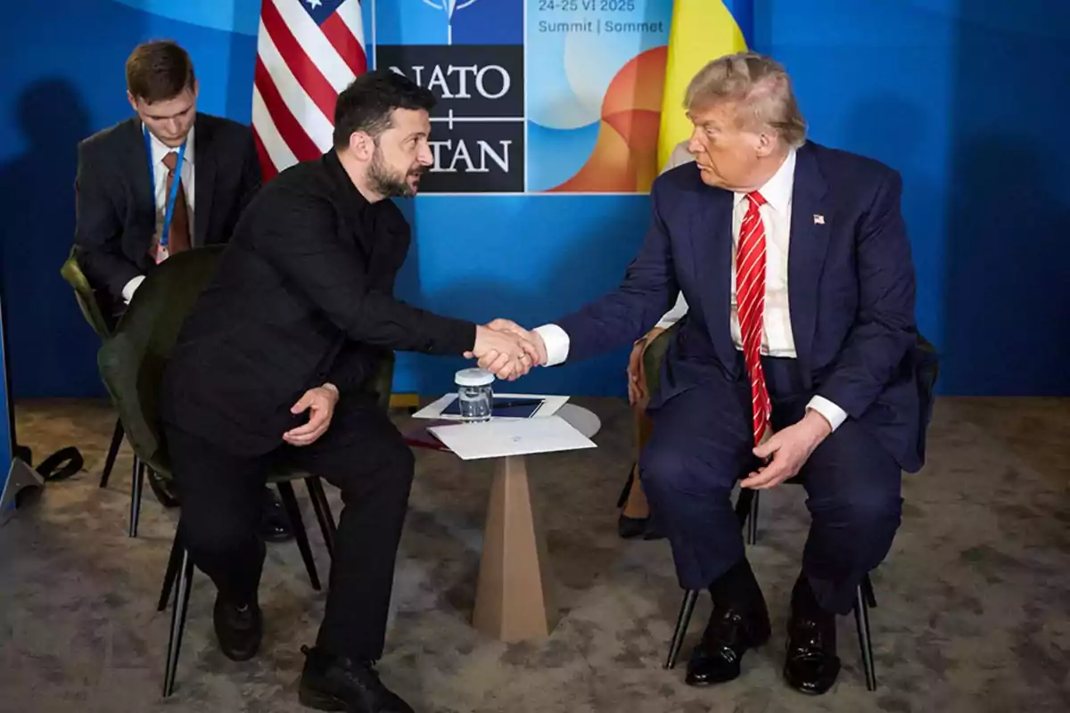 Two men in suits shake hands while seated across a small table during an official meeting with flags in the background and an assistant taking notes. Two men in suits shake hands while seated across a small table during an official meeting with flags in the background and an assistant taking notes.