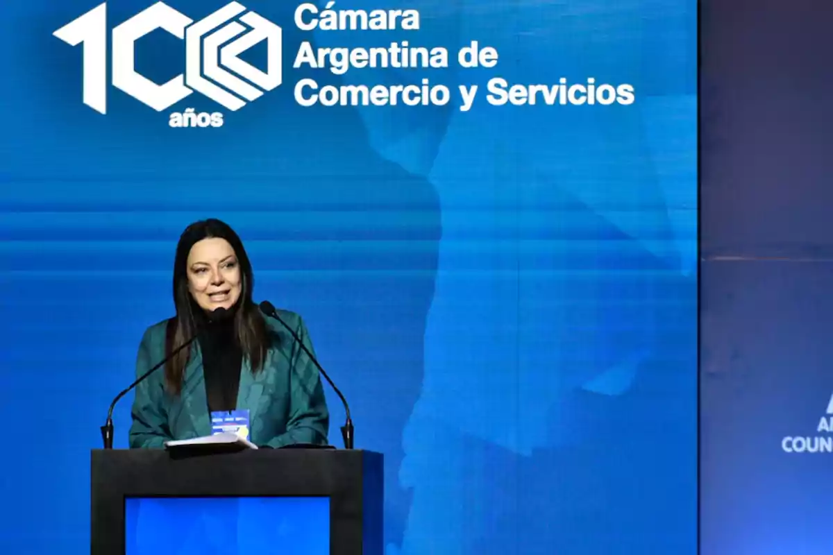 Woman speaking at a podium during an Argentine Chamber of Commerce and Services event with a blue background and institutional logo Woman speaking at a podium during an Argentine Chamber of Commerce and Services event with a blue background and institutional logo
