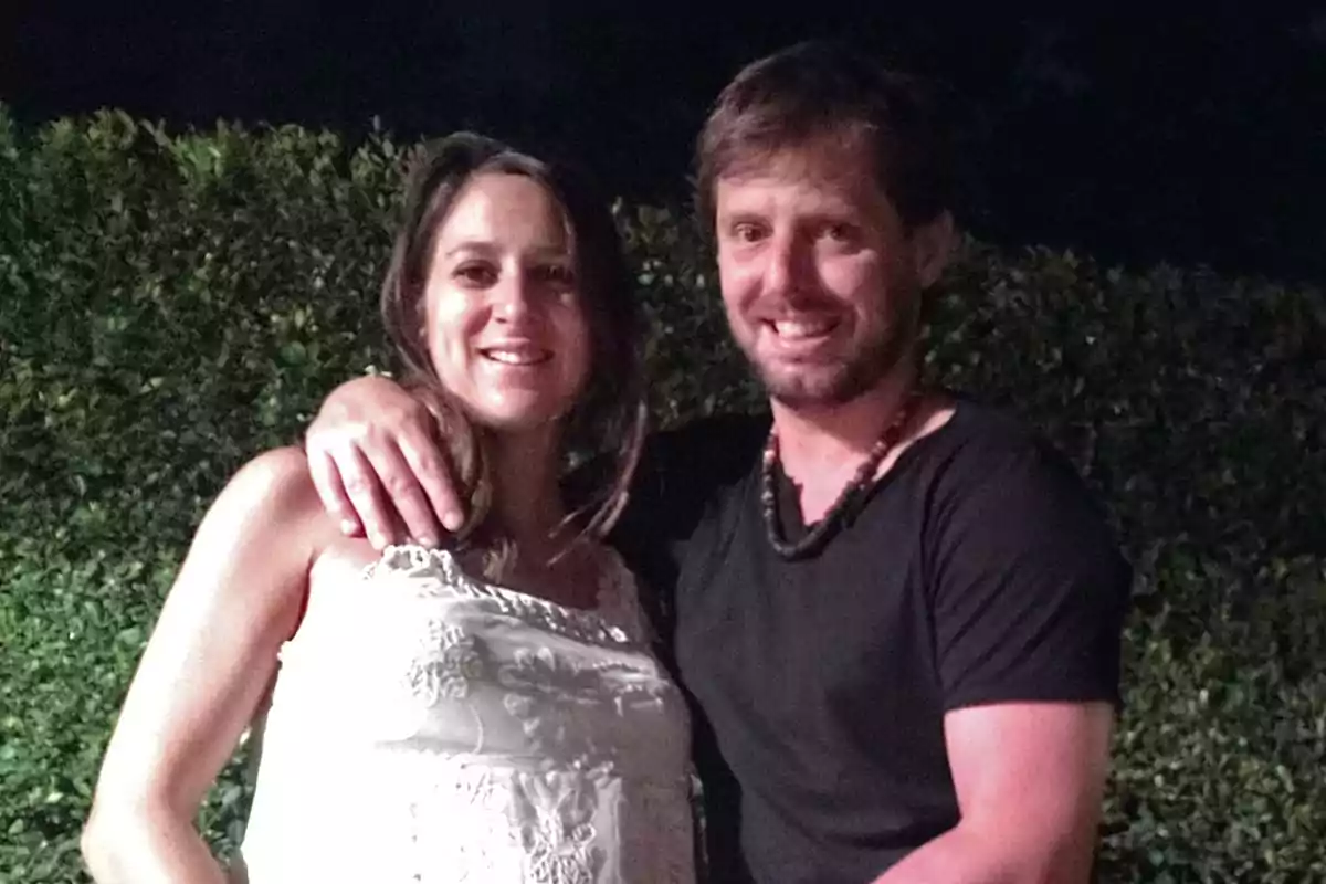 Una pareja sonriente posa abrazada al aire libre frente a un seto verde durante la noche Una pareja sonriente posa abrazada al aire libre frente a un seto verde durante la noche