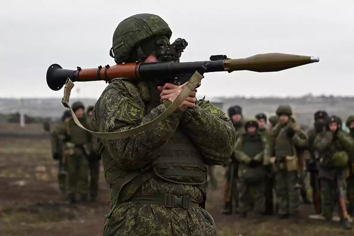 Soldado con uniforme de camuflaje sosteniendo un lanzacohetes mientras otros militares observan al fondo en un campo abierto Soldado con uniforme de camuflaje sosteniendo un lanzacohetes mientras otros militares observan al fondo en un campo abierto