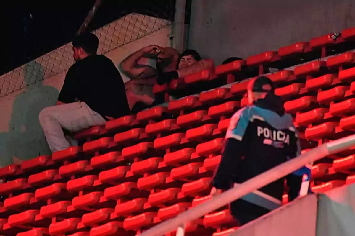 Un policía sube por las gradas de un estadio mientras tres personas están sentadas entre los asientos rojos Un policía sube por las gradas de un estadio mientras tres personas están sentadas entre los asientos rojos