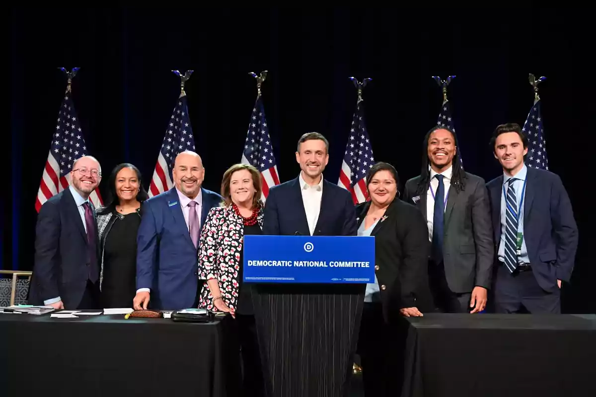 A group of people smiles in front of a Democratic National Committee podium with several American flags in the background. A group of people smiles in front of a Democratic National Committee podium with several American flags in the background.