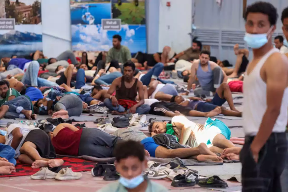 Un grupo numeroso de personas descansa y duerme en el suelo de un espacio cerrado, algunas usando mascarillas y rodeadas de pertenencias y zapatos.