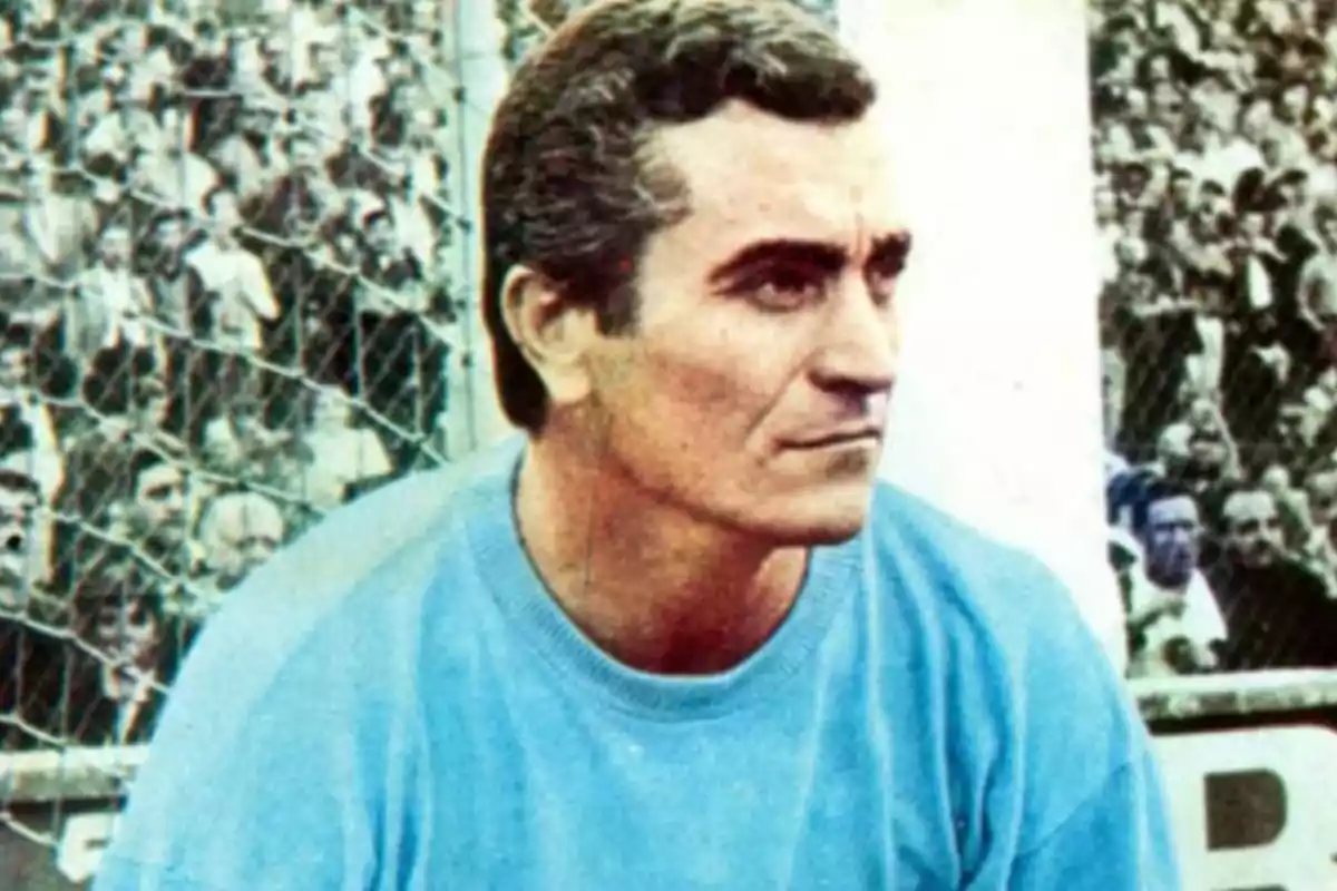 Hombre de cabello corto y canoso vistiendo un suéter azul claro con expresión seria en un estadio lleno de espectadores Hombre de cabello corto y canoso vistiendo un suéter azul claro con expresión seria en un estadio lleno de espectadores