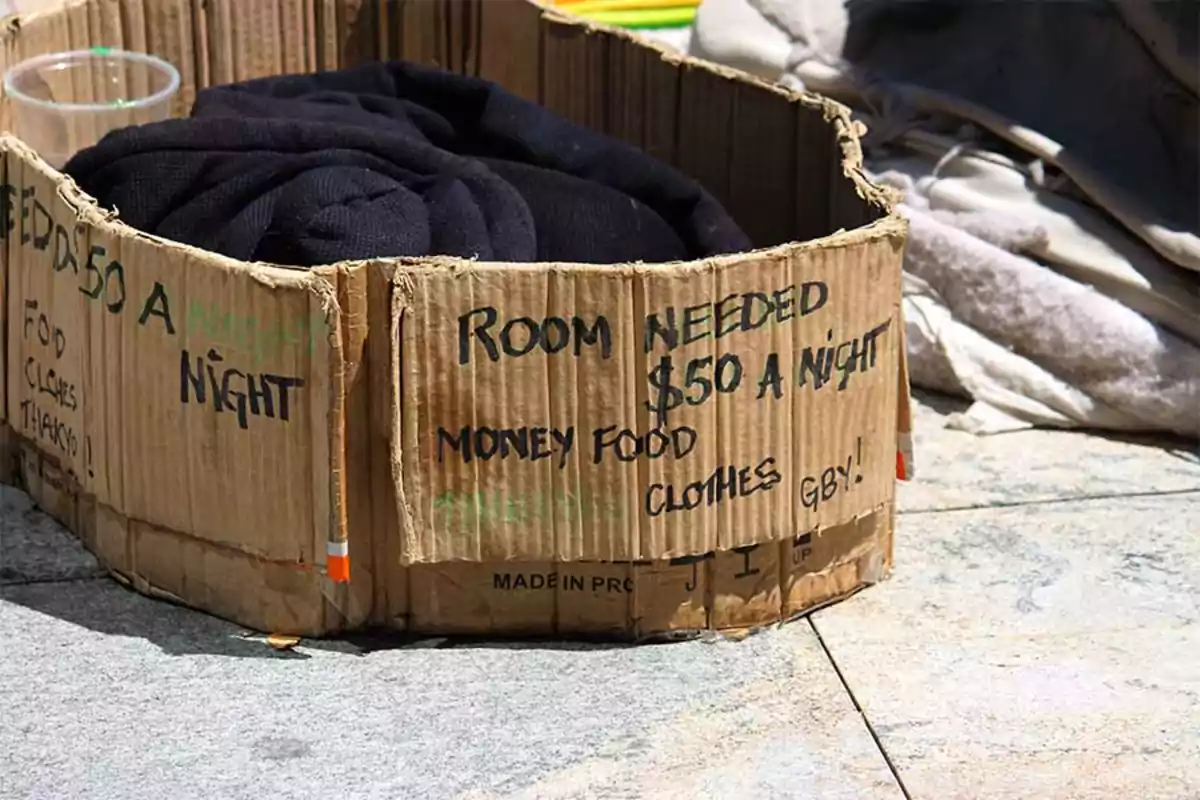 Folded cardboard in a circular shape with a written message asking for help to get money, food, and clothing, next to a dark blanket and some objects on the ground. Folded cardboard in a circular shape with a written message asking for help to get money, food, and clothing, next to a dark blanket and some objects on the ground.