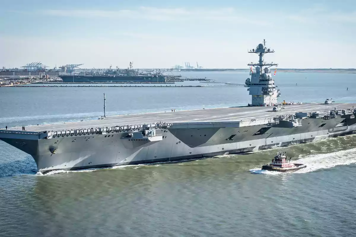 Portaaviones gris navegando en el mar acompañado por un remolcador con un puerto y grúas al fondo Portaaviones gris navegando en el mar acompañado por un remolcador con un puerto y grúas al fondo