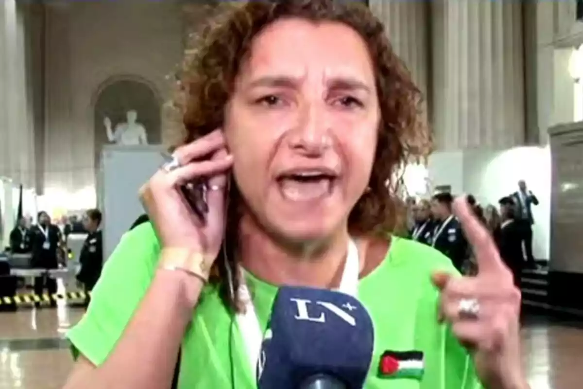 Curly-haired woman in a green t-jersey speaking into a microphone in a building with columns and people in the background Curly-haired woman in a green t-jersey speaking into a microphone in a building with columns and people in the background