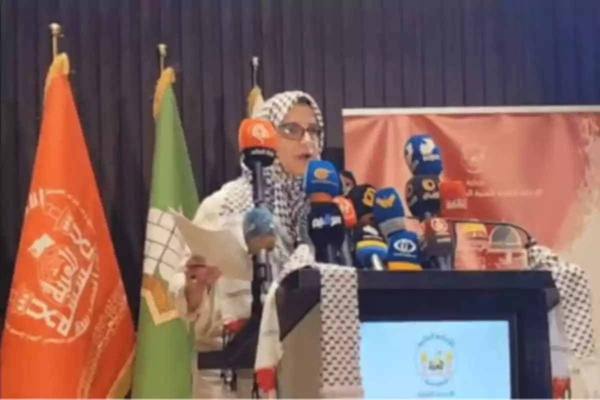 Woman wearing a hijab speaking at a podium surrounded by microphones and flags at an official event