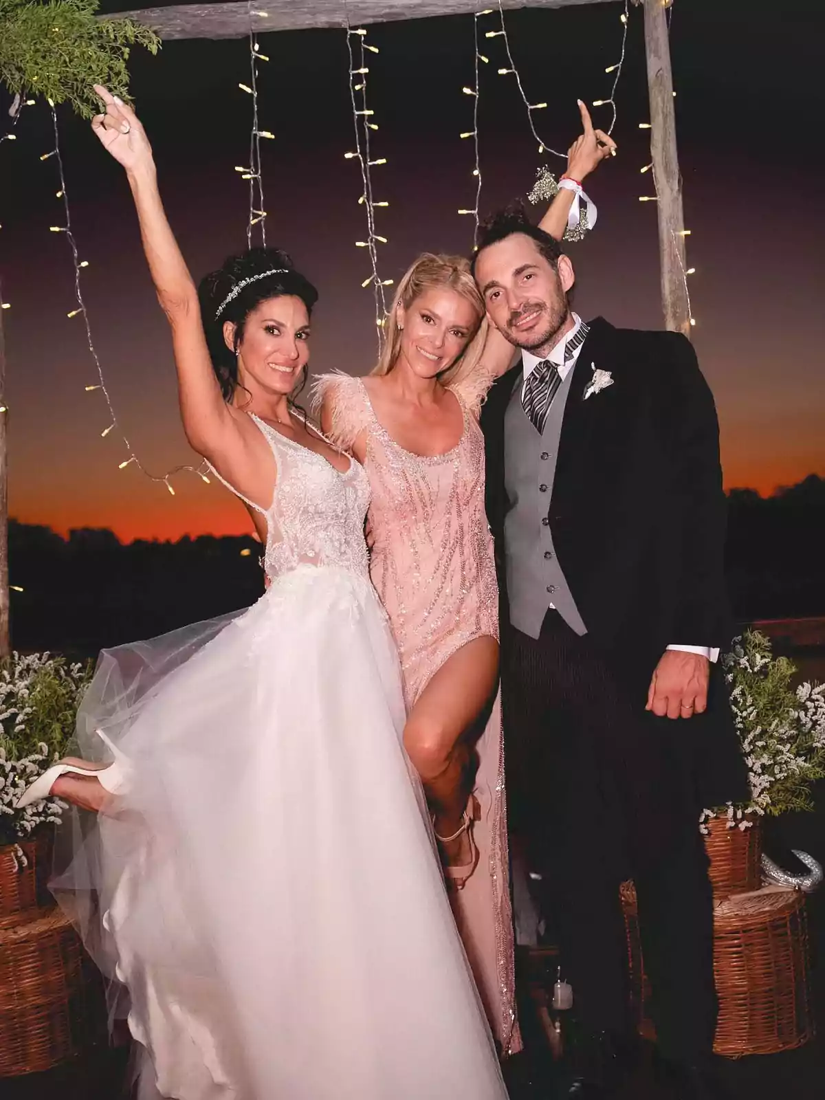 Three people pose smiling at an outdoor wedding at sunset: the bride is wearing a white dress, the groom a dark suit, and the woman in the center a light pink dress; they are all celebrating under decorative lights. Three people pose smiling at an outdoor wedding at sunset: the bride is wearing a white dress, the groom a dark suit, and the woman in the center a light pink dress; they are all celebrating under decorative lights.