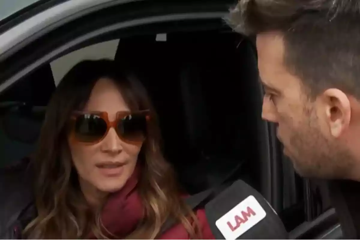 A woman wearing sunglasses is being interviewed by a journalist holding a microphone with the LAM logo while she is sitting in a car.