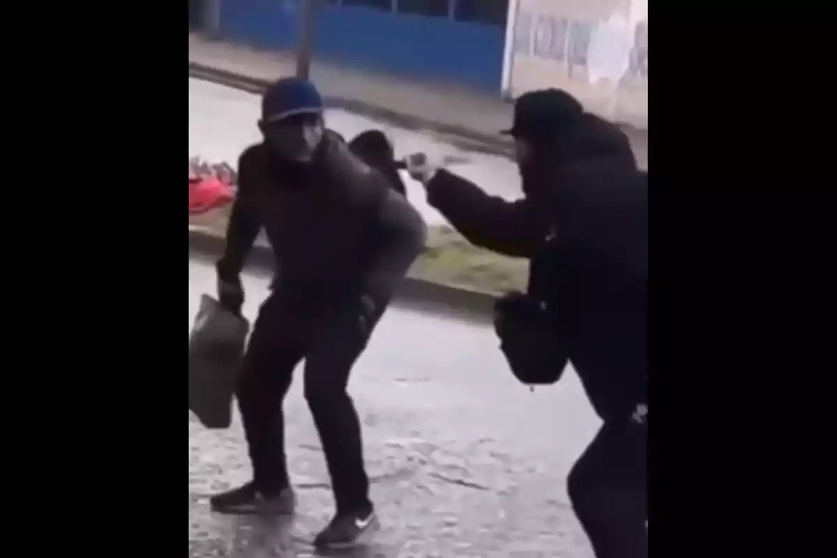 Two people on the street, one of them pointing a gun at the other who is holding a bag and appears to be crouching. Two people on the street, one of them pointing a gun at the other who is holding a bag and appears to be crouching.