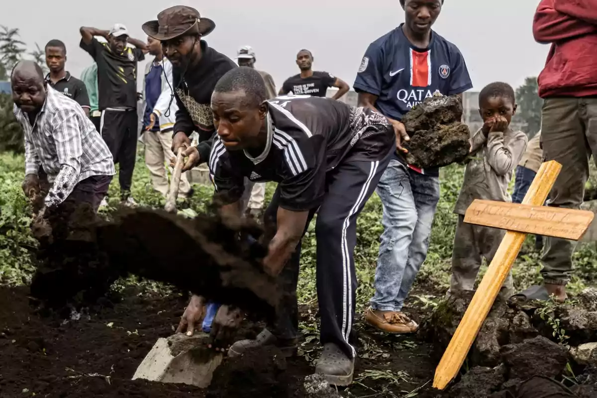 A group of people is burying someone in a field; some are digging while others watch next to a wooden cross.