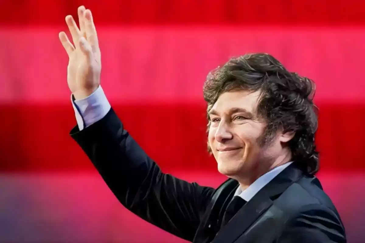 Dark-haired, curly-haired man in a dark suit waving with his hand raised in front of a striped red background.