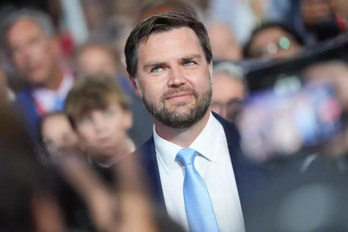 Dark-haired, bearded man in a light blue suit and tie surrounded by people at an event Dark-haired, bearded man in a light blue suit and tie surrounded by people at an event