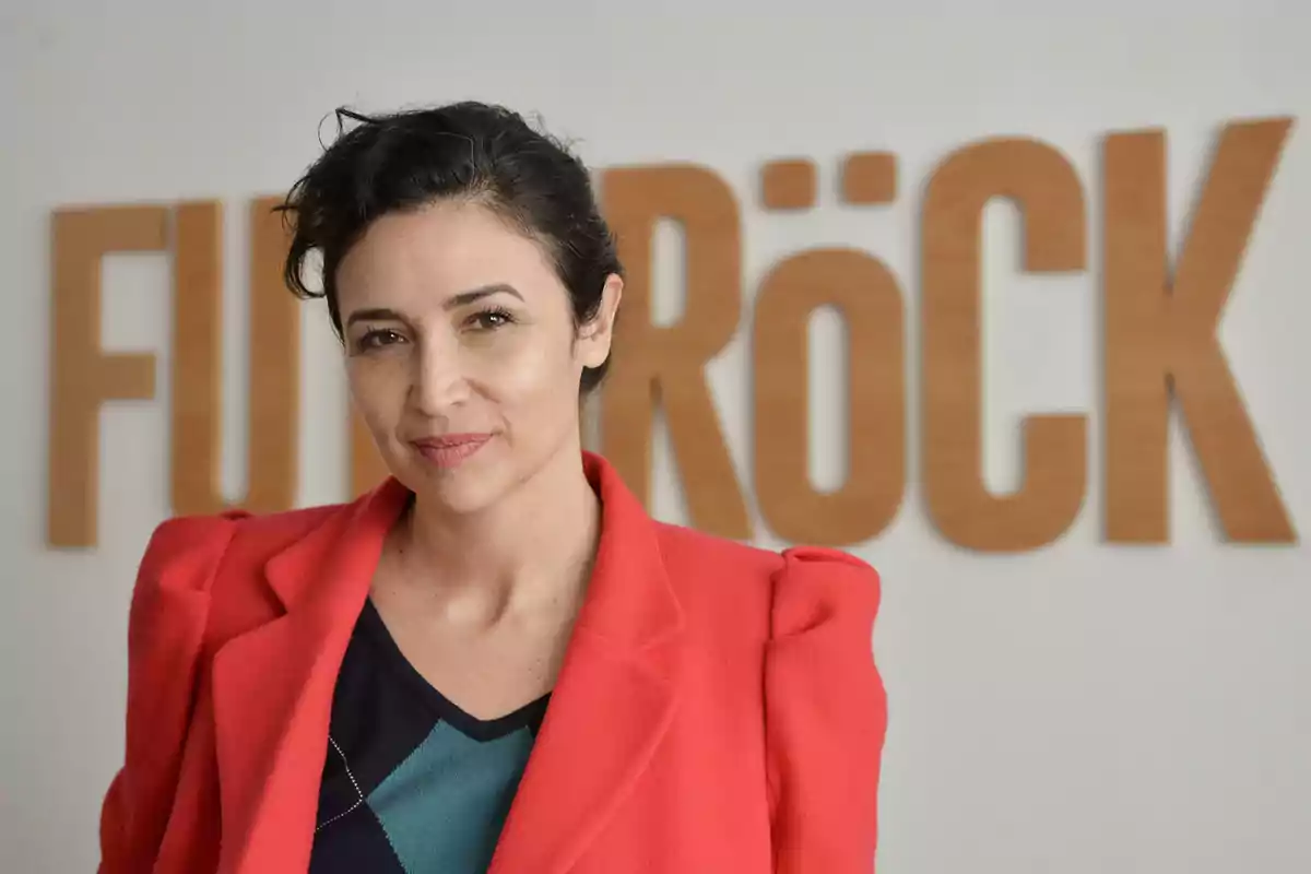Mujer de cabello oscuro y corto con saco rojo posando frente a un letrero de madera que dice FUNRÖCK Mujer de cabello oscuro y corto con saco rojo posando frente a un letrero de madera que dice FUNRÖCK