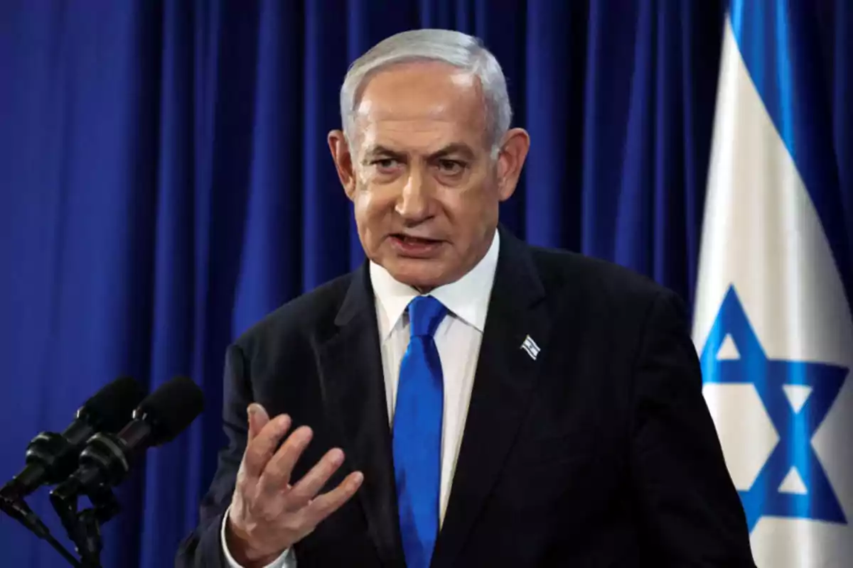 Hombre de cabello canoso con traje oscuro y corbata azul hablando frente a micrófonos con una bandera de Israel al fondo