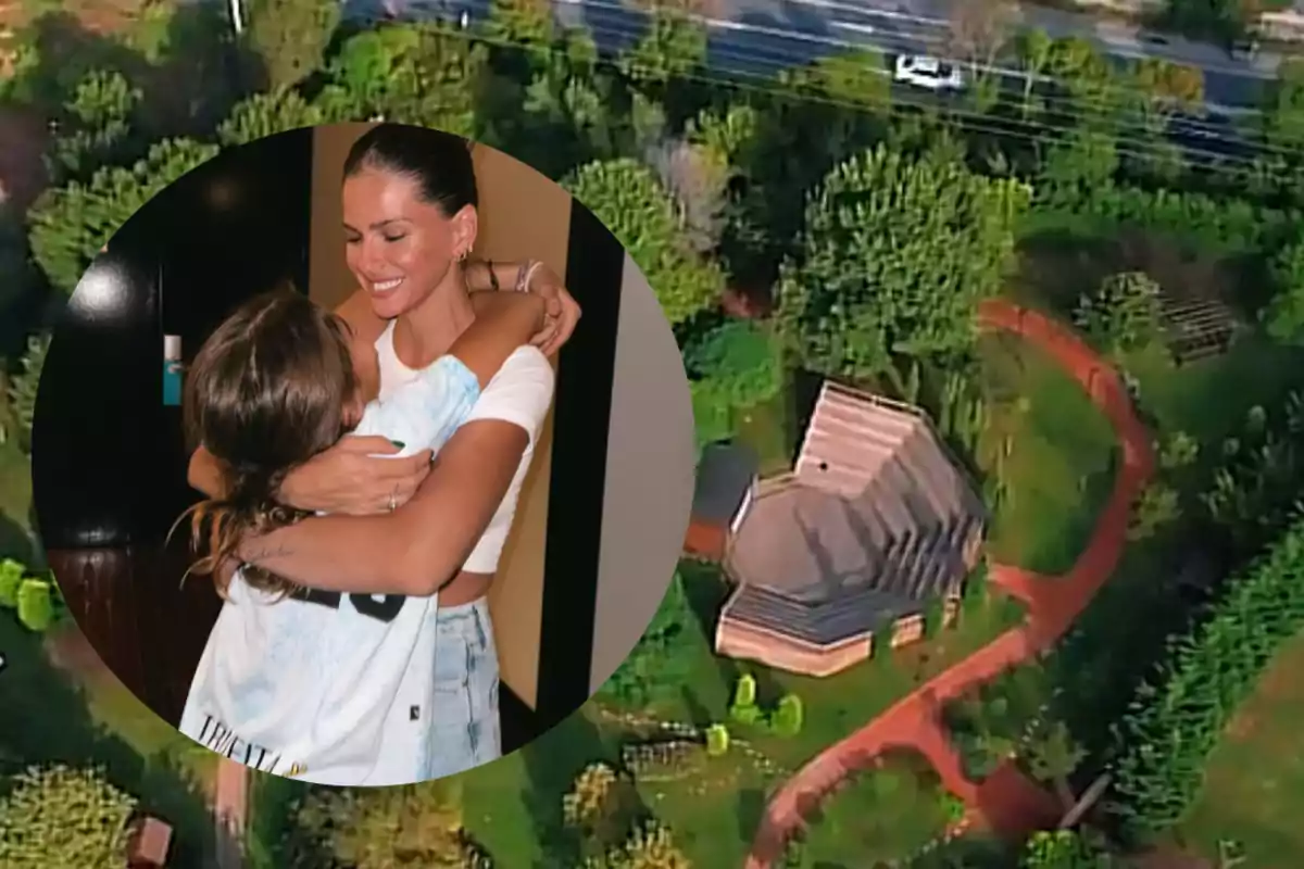 A smiling woman hugs a girl in the foreground while in the background there is a house surrounded by trees and a red path seen from above.