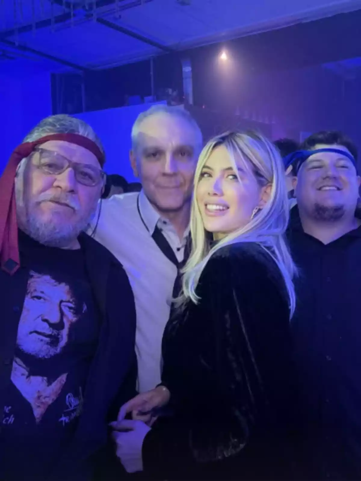 Cuatro personas posan sonrientes en un ambiente de fiesta con luces azules, una de ellas lleva una camiseta con el rostro de un hombre y una cinta roja en la cabeza, mientras que otra persona lleva una cinta azul Cuatro personas posan sonrientes en un ambiente de fiesta con luces azules, una de ellas lleva una camiseta con el rostro de un hombre y una cinta roja en la cabeza, mientras que otra persona lleva una cinta azul