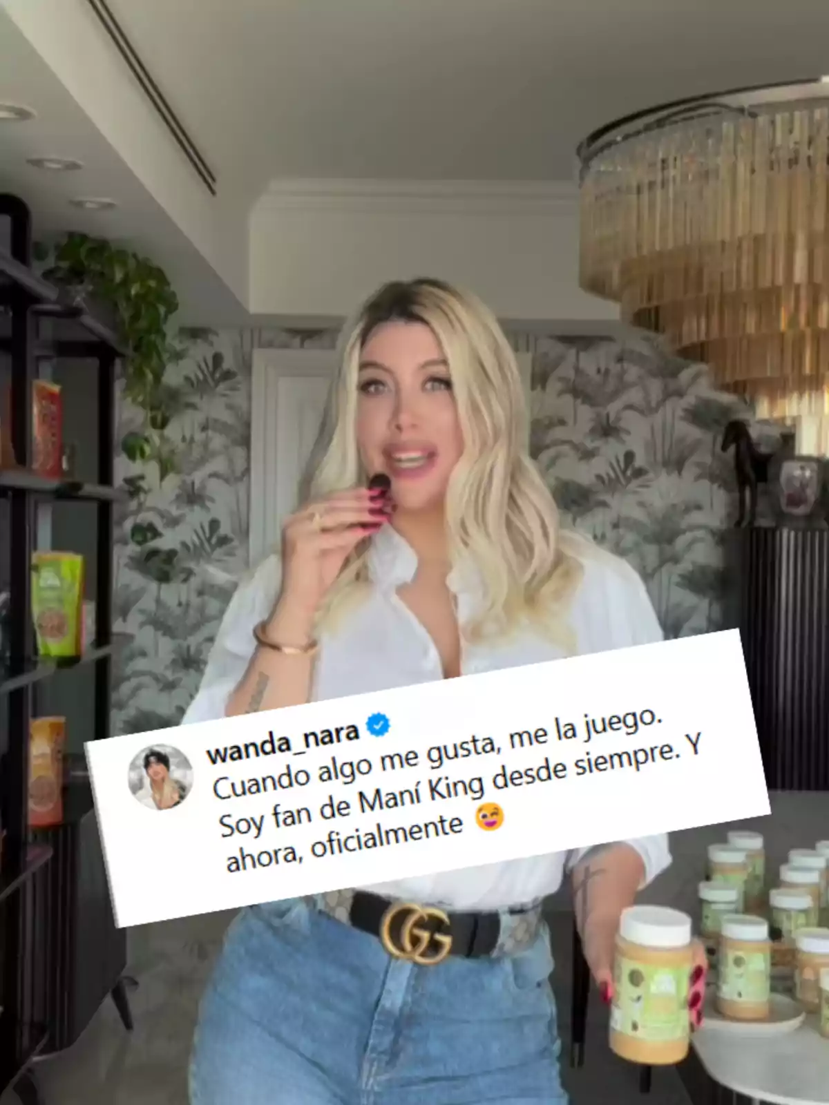 Blonde woman wearing a white jersey and jeans holds a jar of peanut butter in a modern kitchen while holding a food item in her hand, and a box with a promotional message from Maní King appears in the foreground. Blonde woman wearing a white jersey and jeans holds a jar of peanut butter in a modern kitchen while holding a food item in her hand, and a box with a promotional message from Maní King appears in the foreground.