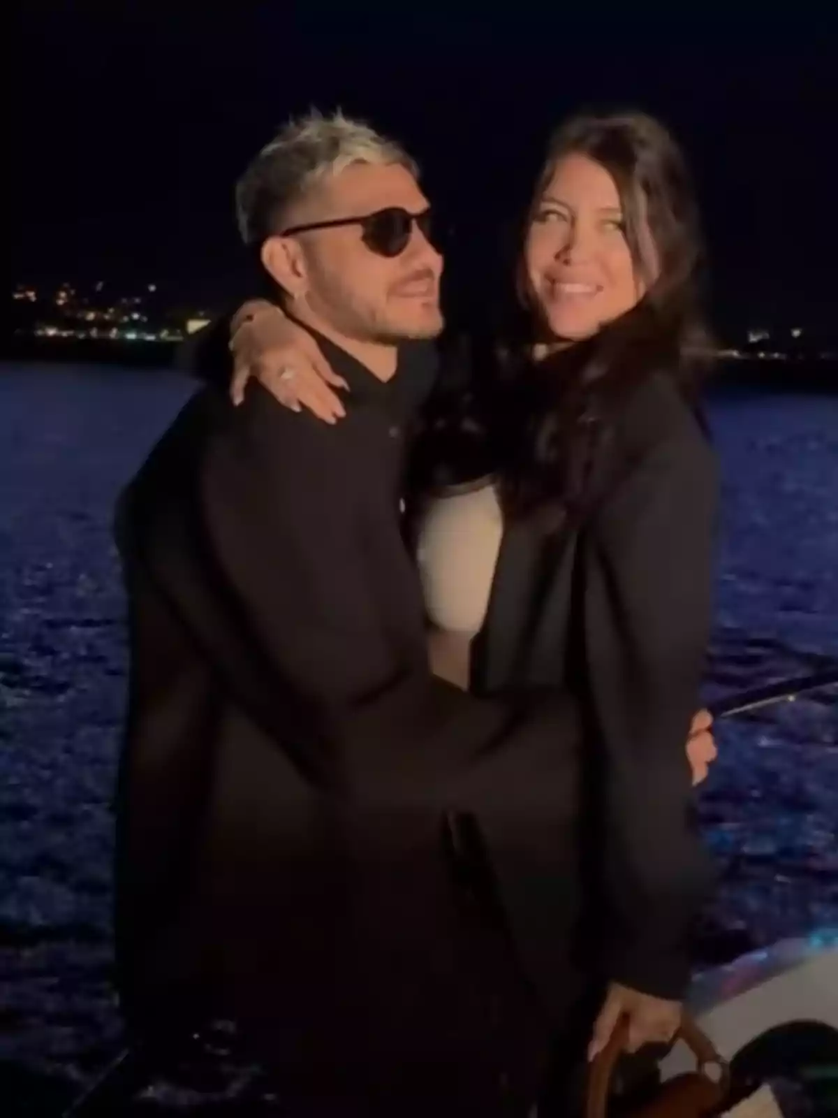 Pareja abrazada en un yate durante la noche con el mar y luces de la ciudad al fondo Pareja abrazada en un yate durante la noche con el mar y luces de la ciudad al fondo