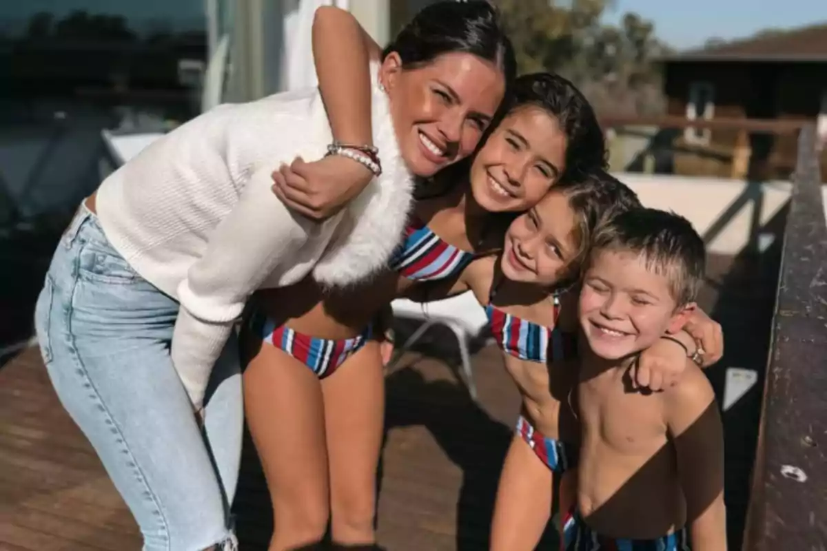 Four people smile, hugging each other outdoors on a sunny terrace: three children in striped swimsuits and a woman in a white sweater and jeans.