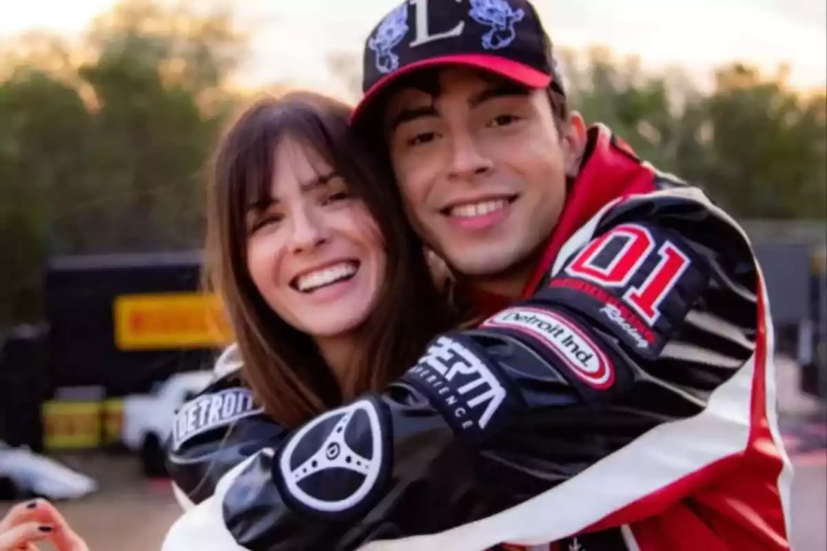 Pareja joven sonriendo y abrazándose con chaquetas de carreras y gorra en un ambiente al aire libre