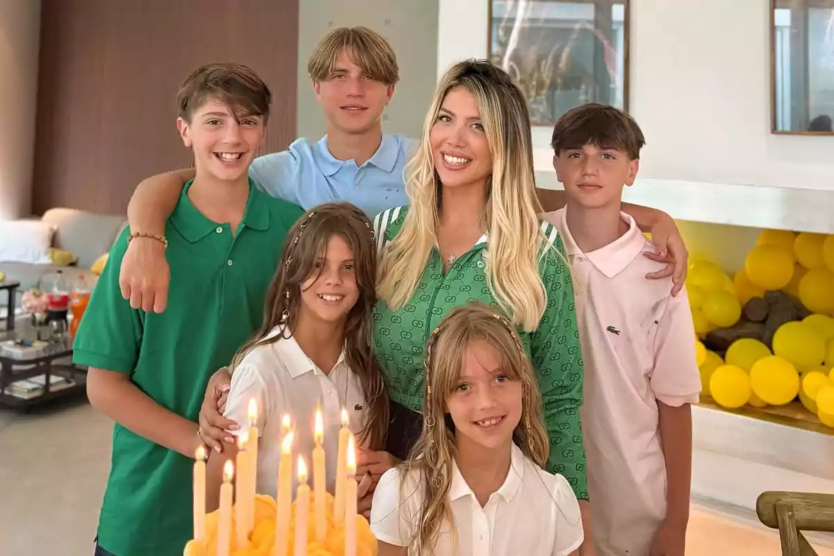 Group of five children and a smiling woman posing together in a room decorated with yellow balloons and a table with a birthday cake with lit candles in front.