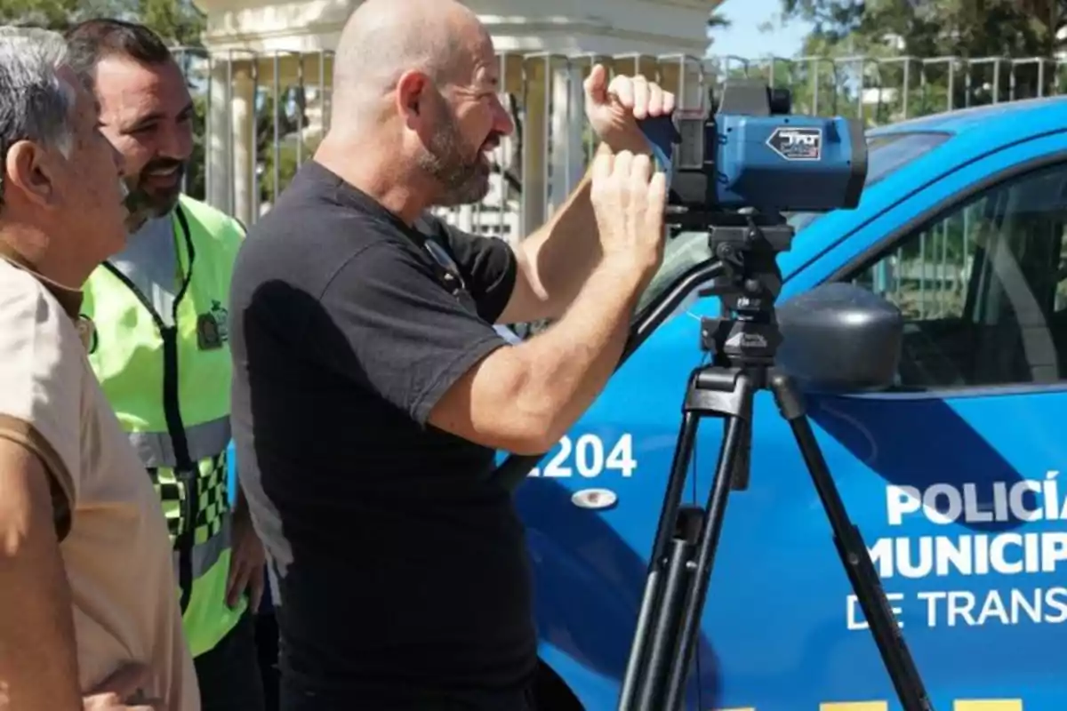 Three men observe a speed radar mounted on a tripod next to a municipal traffic police car.