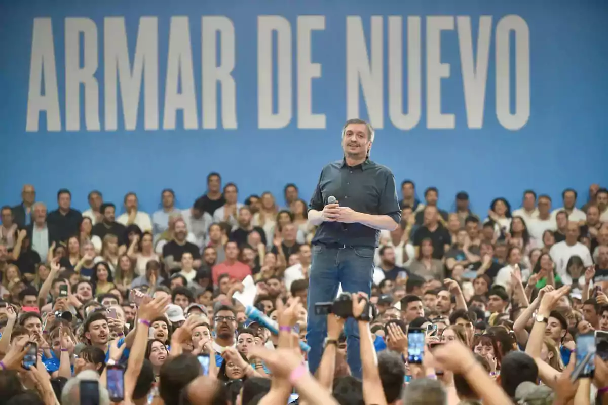 Hombre de pie con micrófono frente a una multitud en un evento con fondo azul y letras grandes que dicen Hombre de pie con micrófono frente a una multitud en un evento con fondo azul y letras grandes que dicen