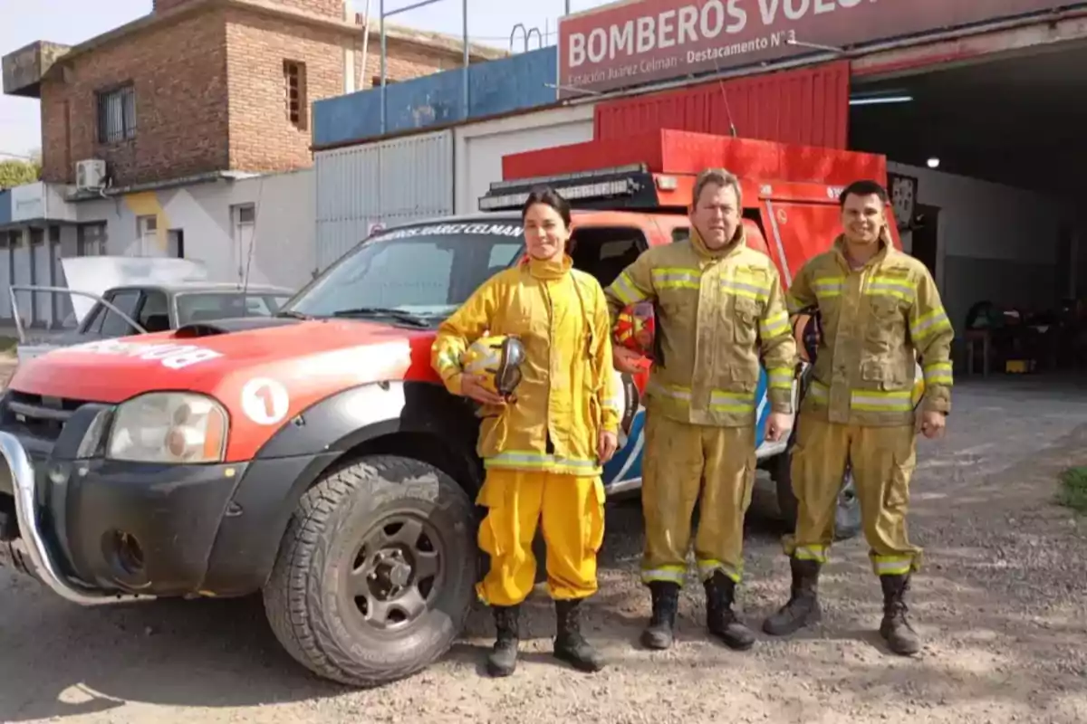 Tres bomberos con uniforme posan frente a una camioneta de bomberos estacionada afuera de una estación Tres bomberos con uniforme posan frente a una camioneta de bomberos estacionada afuera de una estación