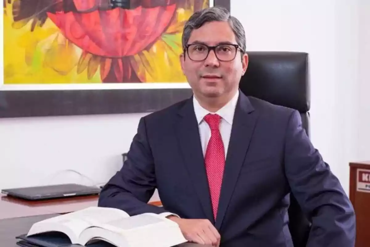 Hombre con gafas y traje oscuro sentado en una oficina con un libro abierto sobre el escritorio y un cuadro colorido en la pared de fondo