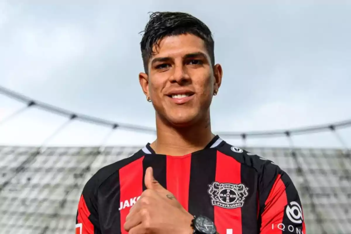 Futbolista con uniforme negro y rojo del Bayer Leverkusen sonríe y muestra el pulgar arriba en un estadio. Futbolista con uniforme negro y rojo del Bayer Leverkusen sonríe y muestra el pulgar arriba en un estadio.